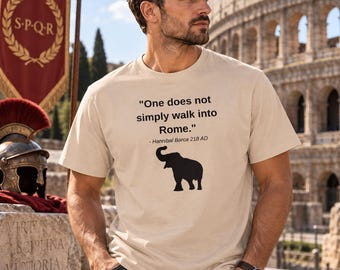 Könnte beinhalten: Ein beiges kurzärmeliges T-Shirt mit schwarzem Text, der "One does not simply walk into Rome." lautet, gefolgt von "- Hannibal Barca 218 AD" und einer schwarzen Elefantensilhouette, die mittig auf der Brust des Kleidungsstücks abgebildet ist.