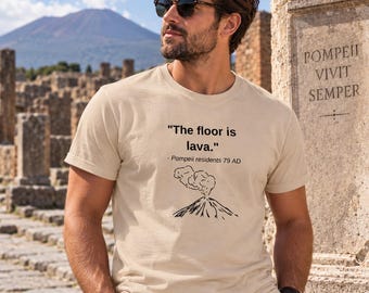 Op de afbeelding: Een beige t-shirt met korte mouwen en zwarte tekst die "The floor is lava." luidt, gevolgd door "- Pompeii residents 79 AD" en een zwarte lijntekening van een uitbarstende vulkaan met rook die uit de top van de berg komt.