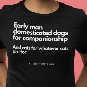 May include: Black t-shirt with white text: "Early man domesticated dogs for companionship. And cats for whatever cats are for." Quote by Philomena Cunk. The shirt is worn with blue jeans against a pink background.