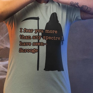 May include: A light green t-shirt with the text "I fear you more than any spectre have seen - Scrooge" and a black Grim Reaper graphic. The person in the image is wearing the shirt and appears shocked.