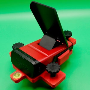 May include: A red and black phone stand with a built-in level. The stand has a black phone holder that can be adjusted to different angles. The stand is made of plastic and has a level on the bottom.