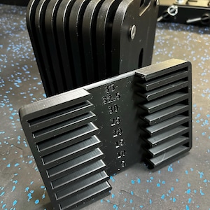 May include: A black plastic weight stack adapter featuring a vertical column of numerical markings from 5 to 35, with horizontal slots on both sides. The device is designed with a series of parallel ridges and a central panel for weight selection.