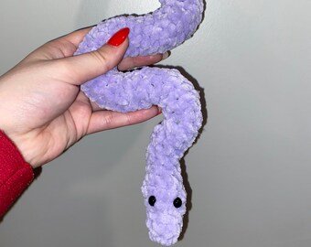 May include: A lavender plush snake toy with black button eyes. The soft, stuffed snake is held against a light grey background. The snake is made of a fuzzy material.