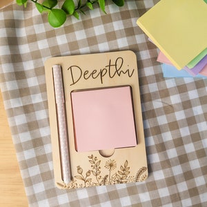 May include: A wooden notepad with the name "Deepthi" engraved on it. The notepad has a pink sticky note and a white pen attached to it. The notepad is decorated with a floral design.