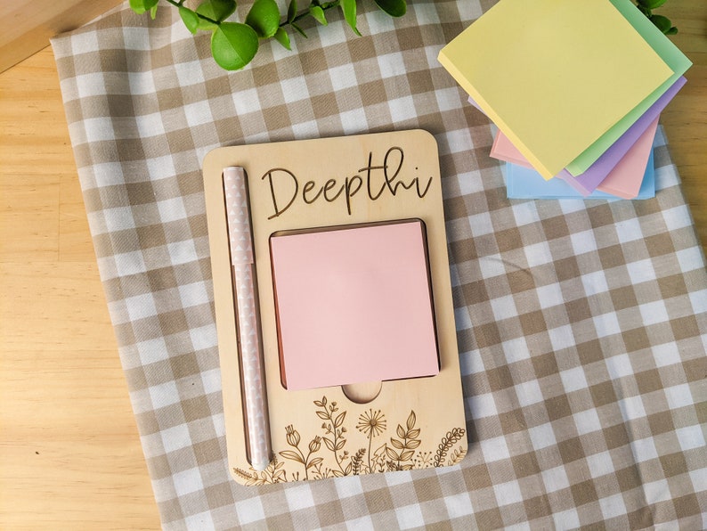 May include: A wooden notepad with the name "Deepthi" engraved on it. The notepad has a pink sticky note and a white pen attached to it. The notepad is decorated with a floral design.