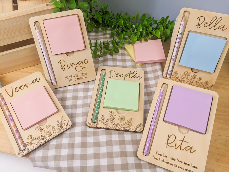 May include: Set of five personalized wooden notepads with a pencil holder. Each notepad has a different name and a floral design. The notepads are pink, light blue, green, purple, and white. The text on the notepads reads "Veena", "Bingo", "Deepthi", "Bella", and "Rita".