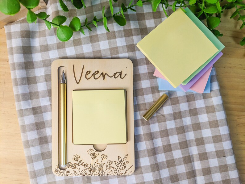 May include: A wooden desk organizer with the name "Veena" engraved on it. The organizer has a slot for a pen and a space for sticky notes. There is a gold pen and a stack of pastel colored sticky notes.