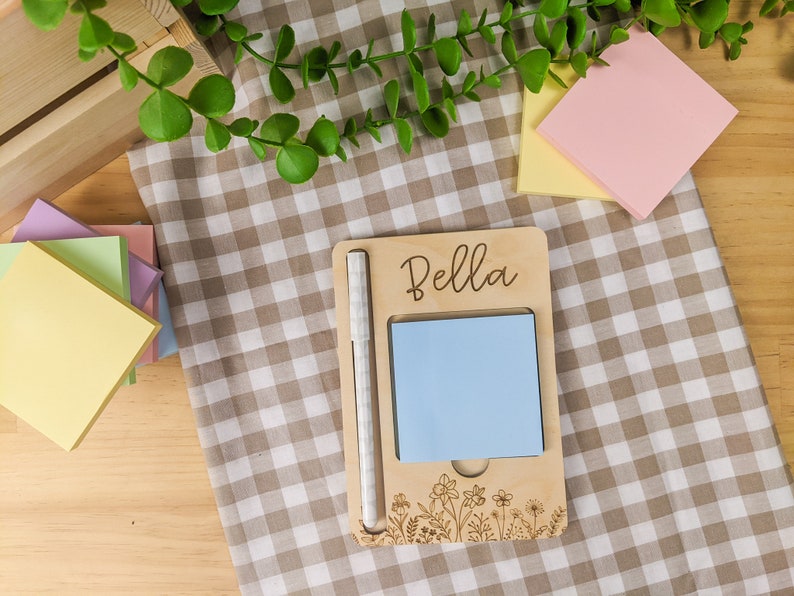 May include: A wooden note holder with a pen, a blue sticky note, and the name "Bella" engraved on the front. The holder has a floral design and is sitting on a checkered tablecloth.