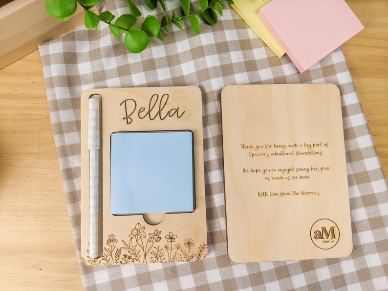 May include: A wooden desk organizer with a light blue sticky note, a white pen, and a floral design. The organizer has the name "Bella" engraved on it and a message thanking the recipient for their support of Spencer's educational foundation. The organizer also has the initials "aM" engraved on it.