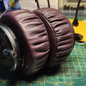 Puede incluir: Un par de auriculares de color morado oscuro con almohadillas negras y un acento plateado. Los auriculares tienen un material similar al cuero que cubre las almohadillas.