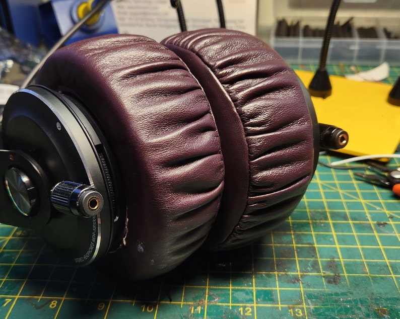 Puede incluir: Un par de auriculares de color morado oscuro con almohadillas negras y un acento plateado. Los auriculares tienen un material similar al cuero que cubre las almohadillas.