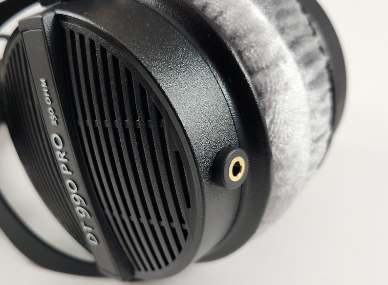 Puede incluir: Auriculares negros con una almohadilla gris y el texto "DT 990 PRO 250 OHM" en el lateral. Los auriculares tienen un conector de audio dorado.
