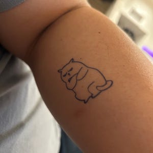 Peut inclure: Un tatouage simple en ligne d'un chat assis, les bras croisés, avec une expression boudeuse.