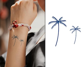 May include: Two palm tree tattoos, one larger than the other, are shown on a person's wrist. The tattoos are a dark blue colour.