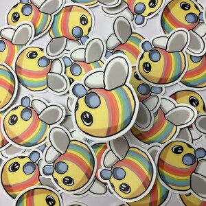 May include: A pile of colorful bee stickers with rainbow stripes and blue eyes. The stickers are on a white background.