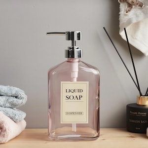 May include: A clear, pink liquid soap dispenser with a silver pump. The label reads "LIQUID SOAP DISPENSER". The dispenser sits on a wooden surface, with folded towels and a fragrance diffuser in the background.
