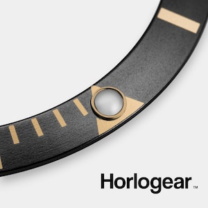 May include: Close-up of a black watch bezel with gold markings and a clear plastic insert. The bezel is curved and has a textured surface. The text "Horologear" is visible in the lower right corner.