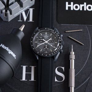 Pu&ograve; includere: Un orologio cronografo Omega Speedmaster nero con cinturino in gomma nero. L'orologio ha una lunetta tachimetrica e un quadrante nero con indicazioni bianche. L'orologio &egrave; su una superficie nera con uno strumento e altri accessori per l'orologeria.