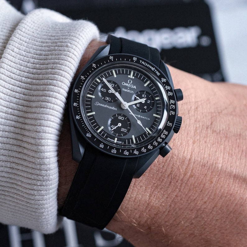 Pu&ograve; includere: Un orologio cronografo Omega Speedmaster nero con cinturino in gomma nero. Il quadrante dell'orologio ha una scala tachimetrica e le parole "Omega swatch" stampate su di esso.