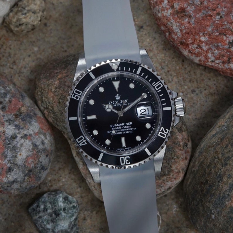 Op de afbeelding: Een zwart Rolex Submariner horloge met een witte bezel en een witte rubberen band. De wijzerplaat van het horloge heeft een zwarte achtergrond met witte markeringen en een datumvenster op 3 uur. Het horloge ligt op een grijze achtergrond.