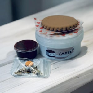 May include: A white plastic container with a red heart pattern lid, filled with a brown cookie and labeled "S'MORE". A small clear plastic bag with brown and gold sprinkles and a small red and white container with a dark red liquid are also shown.