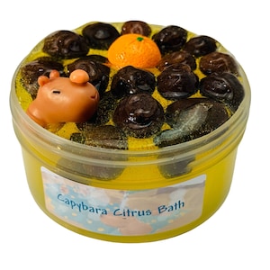 Op de afbeelding: Een gele slijm met een capybara-figuur en sinaasappelschijfjes in een doorzichtige plastic container. De container heeft een label met de tekst "Capybara Citrus Bath".