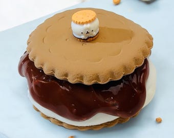May include: A chocolate-filled s'more with a marshmallow and two graham cracker cookies. A small, brown and white character is on top of the top cookie.