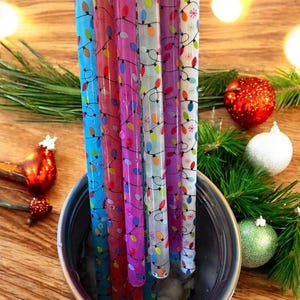 Peut inclure: Gros plan sur des pailles colorées à motifs dans un récipient en métal. Les pailles sont décorées de guirlandes lumineuses de Noël de différentes couleurs. Des décorations de Noël et des branches de pin sont en arrière-plan.