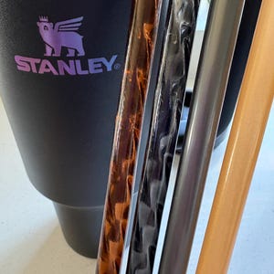 Op de afbeelding: Close-up van vier verschillende gekleurde drinkrietjes. De rietjes zijn verticaal gerangschikt en bevinden zich voor een zwarte Stanley-reismok met een paars-blauwe gradiëntdeksel. De mok heeft een logo van een gevleugeld wezen met een kroon op zijn hoofd en het woord "STANLEY" eronder.