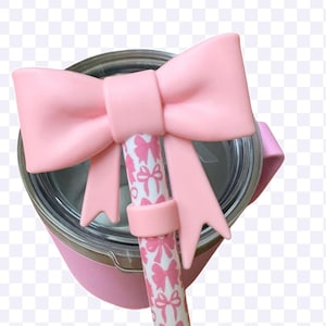 以下が含まれることがあります： ピンク色のタンブラーと、お揃いのピンク色のリボン型のストローのトッパー。ストローは、ピンクのリボン模様の白い背景です。タンブラーもピンク色で、ハンドルが付いています。