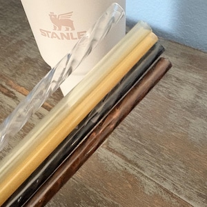 May include: Four reusable drinking straws in clear, translucent gold, translucent black, and brown wood-grain patterns. The clear straw features a twisted spiral design, while the others are smooth. A light pink Stanley tumbler is visible behind them.