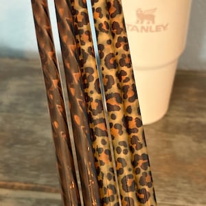 May include: Four cylindrical drinking straws arranged vertically. Two straws feature a dark brown, translucent, swirling pattern, while the other two straws display a repeating black and orange leopard print design against a translucent base.