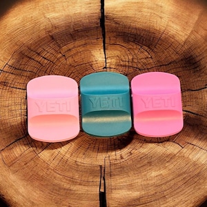 May include: Three Yeti drinkware accessories in pink and teal. Each has the Yeti logo embossed on the front. The accessories are displayed on a wooden surface with a natural grain pattern.