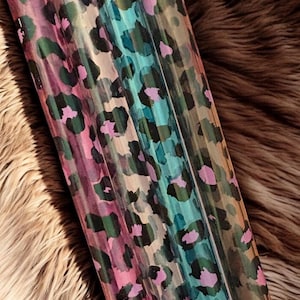 Könnte beinhalten: Eine Rolle gemusterter Vinylfolie mit Leopardenmuster. Der Druck enthält Farbtöne von Rosa, Grün, Blau und Beige. Das Vinyl wird vor einem Hintergrund aus Kunstfell gezeigt.