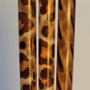 May include: Three cylindrical rods arranged vertically. The two rods on the left feature a brown and tan leopard print pattern. The rod on the right displays a translucent amber and brown marbled pattern with a smooth, glossy finish.