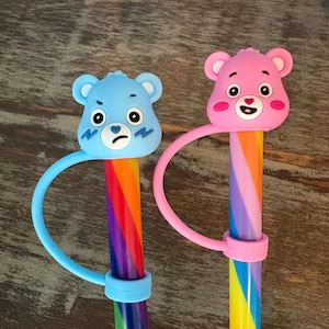 Könnte beinhalten: Zwei Stifte mit regenbogenfarben gestreiften Schäften und Silikonaufsätzen in Form von Bärenköpfen. Ein Aufsatz ist hellblau mit mürrischem Gesichtsausdruck, der andere ist rosa mit lächelndem Gesicht, jeweils befestigt an einer passenden Silikonschlaufe.