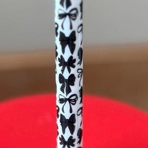 May include: A white reusable drinking straw featuring a repeating pattern of small black bows. The straw is inserted into the centre of a bright red circular lid, which is part of a larger container not fully visible in the frame.