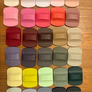 Puede incluir: Una colección de treinta abridores de botellas rectangulares de silicona dispuestos en una cuadrícula, cada uno con el logotipo YETI en relieve. Los artículos se presentan en una gran variedad de colores sólidos, incluyendo blanco, rosa, rojo, marrón, beige, azul, verde, amarillo, gris y negro.