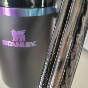 May include: Black Stanley tumbler with a purple and iridescent trim. The Stanley logo is in purple. A black, glittery straw is in the foreground. The tumbler is designed to keep drinks cold or hot for hours.