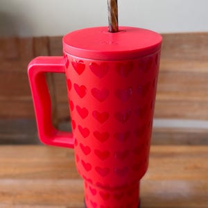 May include: Red travel mug with a heart pattern and a black lid. The mug has a handle and a straw.