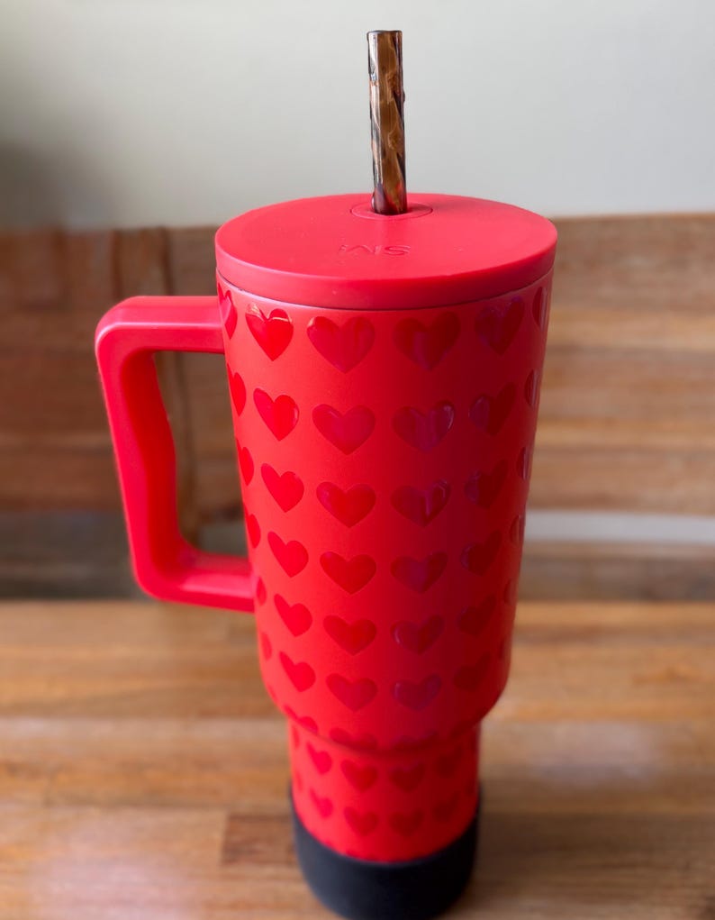 May include: Red travel mug with a heart pattern and a black lid. The mug has a handle and a straw.
