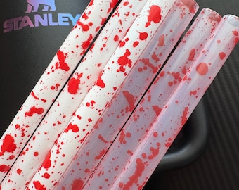 Può includere: Cannucce di plastica trasparente con base bianca e design a spruzzi rossi. Le cannucce sono angolate nell'immagine, con un bicchiere Stanley nero sullo sfondo. Il logo Stanley è visibile.