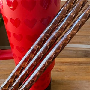 May include: Three brown and gold swirled drinking straws with a red heart patterned tumbler in the background.