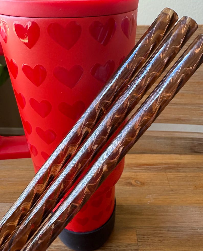 May include: Three brown and gold swirled drinking straws with a red heart patterned tumbler in the background.