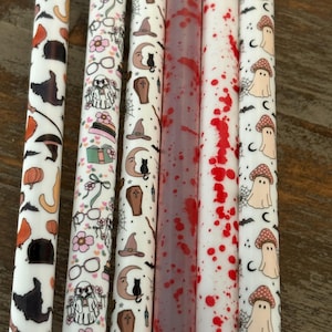 May include: A set of six cylindrical straws featuring various patterns including orange pumpkins, black cats on crescent moons, coffins, witch hats, books, glasses, flowers, red blood splatter, and beige ghosts with spotted mushroom caps on a white background.