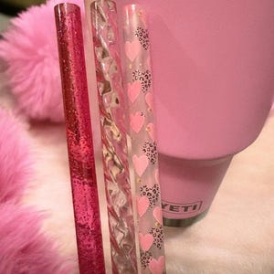 May include: Three decorative drinking straws. One is a solid pink with glitter, the second is clear with pink swirls, and the third is clear with pink hearts and leopard print accents. A pink tumbler is in the background.