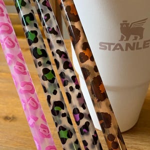 Op de afbeelding: Vier doorzichtige rietjes met luipaardprint in roze, groen, paars en bruin. De rietjes staan voor een witte Stanley-beker met het merklogo.