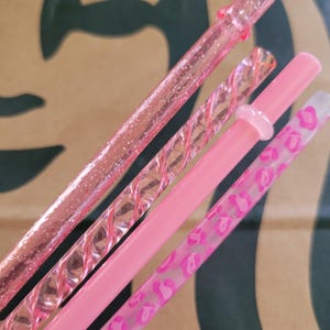 May include: Four pink reusable straws. One is glittery, one has a twisted design, one is solid, and the last has a pink leopard print pattern. The straws are angled against a brown and green background.