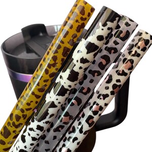 May include: Four reusable straws with animal print designs, including leopard and cow patterns, are displayed. The straws are yellow, black, white, and gray, with pink accents. A black tumbler is partially visible in the background.