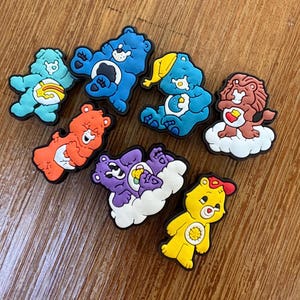 Peut inclure: Une collection de breloques de chaussures Care Bears colorées. Les breloques présentent divers Care Bears dans des tons de bleu, orange, jaune, violet et marron, chacun avec des motifs et des expressions uniques. Ils sont posés sur un fond en bois.